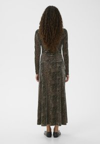 Frau mit langen lockigen Haaren, die ein langes, ärmelloses, knöchellanges Kleid mit Leopardenmuster und schwarze flache Schuhe trägt und mit dem Rücken zur Kamera steht.