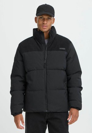 Schwarze Pufferjacke mit einem kontrastierenden strukturierten oberen Abschnitt, Reißverschluss und elastischen Bündchen, getragen über einem grauen T-Shirt und schwarzen Hosen.