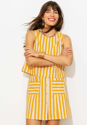 Femme souriante aux cheveux bruns portant un haut sans manches rayé jaune et blanc et une jupe assortie avec des poches devant, les bras croisés.