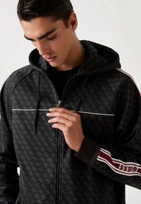 Felpa nera con zip, con una trama decorativa, dettagli bianchi e strisce bordeaux in evidenza sulle maniche. Include un cappuccio e tasche frontali.