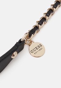 Musta nahkaranneke, jossa on kultainen metalliketju ja pyöreä tunnus, jossa lukee "GUESS EST.1981." Sileä pinta, jossa kiiltävä viimeistely metalliosissa.