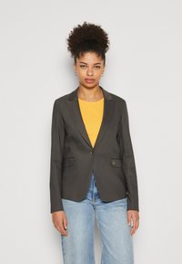 Tmavě šedý upravený blazer s jedním knoflíkem, zářezovými klopami, dvěma předními kapsami a hladkým povrchem, nošený přes žlutý top.