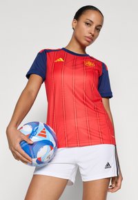 Woman in red and navy Spain soccer jersey and white Adidas shorts holding a blue and white soccer ball by her side.