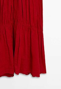 Punainen, porrastettu mekko, joka on valmistettu kevyestä, teksturoidusta kankaasta. Mekossa on kerätty vyötärö ja virtaava helma, jossa on pieni aalto alaosassa.