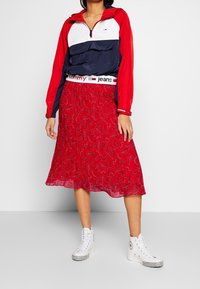 Roter Paisley-Rock mit schwingendem Design, kombiniert mit einer navyblauen, weißen und roten Halbreißverschluss-Windjacke und weißen High-Top-Sneakern.