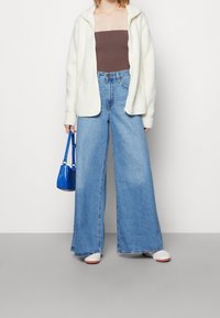 Vit fleece zip-jacka, brun bandeau topp, vida ljusblå jeans, vita sneakers och en liten blå handväska. Släta texturer, avslappnad passform.