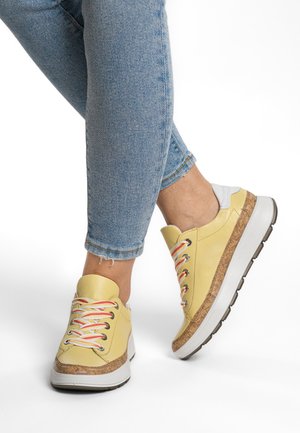 Yellow leather sneakers with a cork base, multicolored laces, and a white accent at the heel, paired with rolled denim jeans.