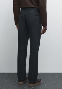 Pantalon noir à coupe ajustée, en tissu lisse. Dispose de poches latérales et d'une poche arrière discrète. Porté avec des chaussures marron.