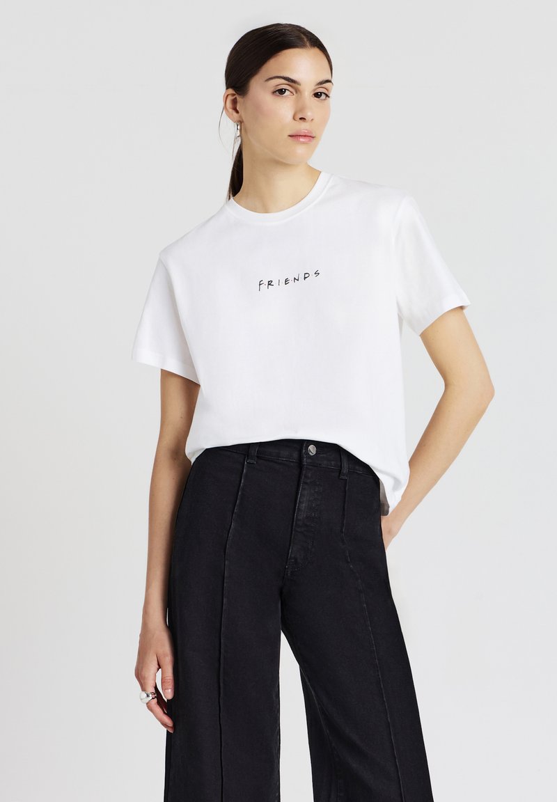 T-shirt bianca in cotone con collo tondo e maniche corte, che presenta la parola "FRIENDS" in lettere nere sul petto. Abbinata a pantaloni neri a vita alta.