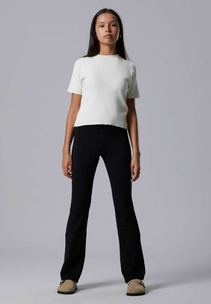 Nainen seisoo harmaata taustaa vasten pukeutuneena valkoiseen lyhythihaiseen puseroon, mustiin housuihin ja ruskeisiin slip-on-kenkiin, katsoo suoraan eteenpäin neutraali ilme kasvoillaan.