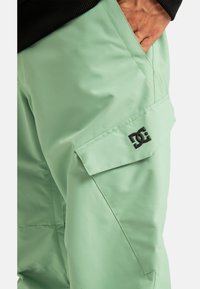 Zielone wodoszczelne spodnie posiadają boczną kieszeń cargo oraz czarne logo. Materiał jest gładki i nieco błyszczący, a krój jest luźny.