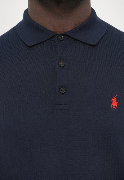 Tengerészkék pólóing bordázott anyagból, klasszikus gallérral, három fekete gombbal és piros hímzett logóval a mellkason.