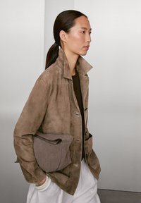 Bruine suede jas met een hoge kraag, voorzakken en knoopsluiting. Houdt een zachte, grijze handtas vast. Model met een paardenstaart.
