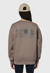 Brun sweatshirt med rund hals, med turkost tryckt text och grafik på ryggen. Matchas med en beige keps.