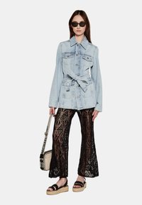 Giubbotto di jeans azzurro chiaro con vita piatta, ampie tasche e dettagli con bottoni, abbinato a pantaloni ampi a maglia nera con texture.