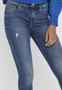 Vaqueros de denim azul con un corte ajustado, que presentan un parche desgastado en el muslo, cinco bolsillos y costuras en contraste. La textura es suave y ligeramente desvaída.