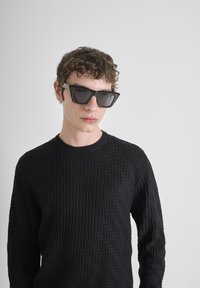 Zwarte trui met een gestructureerd visgraatpatroon en hoekige zwarte zonnebril met donkere lenzen, gedragen door een persoon met krullend haar.