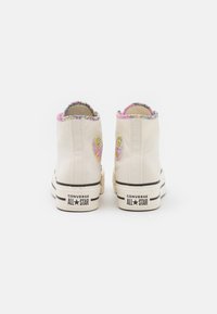 Helle Canvas-High-Top-Sneaker in Creme mit pinken Blumenakzenten, verziert mit Herz-Patches, ausgestattet mit einem schwarzen Streifen und "Converse All Star" Branding.