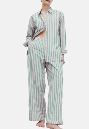 Persona che indossa un pigiama largo a righe verticali rosa e verdi, con maniche lunghe e pantaloni a gamba larga, con una mano in tasca.