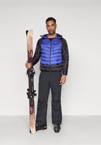 Casaco puffer azul e preto, calças de esqui pretas e botas de esqui pretas. Segura esquis de madeira com fixações; aderência sólida, base plana.