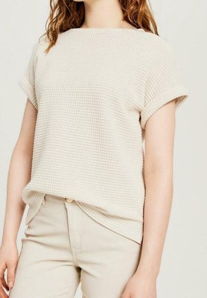 Vrouw die een crèmekleurige, textuurvolle top met korte mouwen draagt, ingestopt in een beige broek met hoge taille, staand tegen een effen achtergrond.