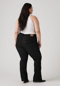 Black denim flared jeans with back pockets, high waistband, and leather brand patch. Model wears a white tank top. Smooth texture.