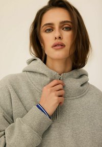 Szara bluza z kapturem na zamek, z wysokim kołnierzem, prążkowanymi mankietami i wypukłym logo. Posiada miękką teksturę oraz niebieski prążkowany akcent na nadgarstku.