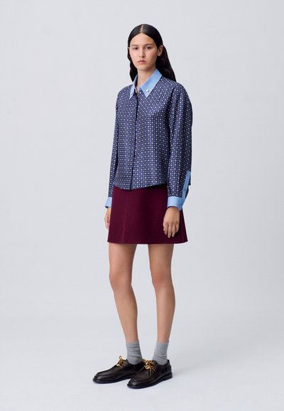 Camisa de patrones marinos con cuello azul claro, falda burdeos, calcetines grises y zapatos negros con cordones dorados. Diseño de corte recto y detalles clásicos.