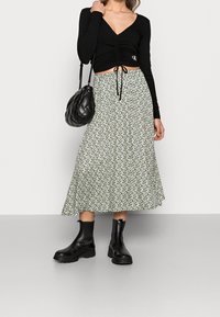 Svart räfflad långärmad topp med samlad detaljer, parat med en grön blommig midiklänning. Modellen håller en svart fyllig väska och har på sig svarta stövlar.