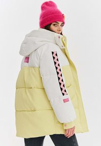 Pufová bunda v bielej a pastelovo žltej farbe, s kapucňou, fialovými a ružovými akcentmi a zvlneným ružovým vzorom na rukáve.