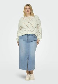 Jersey de punto crema con patrón de diamantes, combinado con una falda maxi de mezclilla azul claro con abertura frontal y zapatos planos beige.