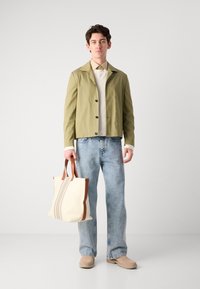 Olijfgroene lichtgewicht jas met zwarte knopen, crèmekleurige sweatshirt, lichtblauwe denim jeans en een crèmekleurige tote bag met bruine handvatten.