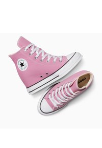 Des baskets montantes en toile rose avec des œillets et des lacets blancs, des semelles en caoutchouc noires et un patch rond avec le logo sur le côté extérieur.