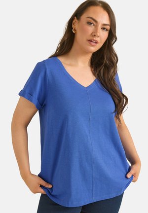 Kvinde med langt brunt hår iført en blå kortærmet V-hals T-shirt, holder i siderne af trøjen og poserer foran en ensfarvet baggrund.