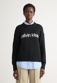 Schwarzer Strickpullover mit "Calvin Klein" Logo, Rundhalsausschnitt, überschnittenen Schultern, kombiniert mit einem hellblauen gestreiften Hemd und beigen Hosen.
