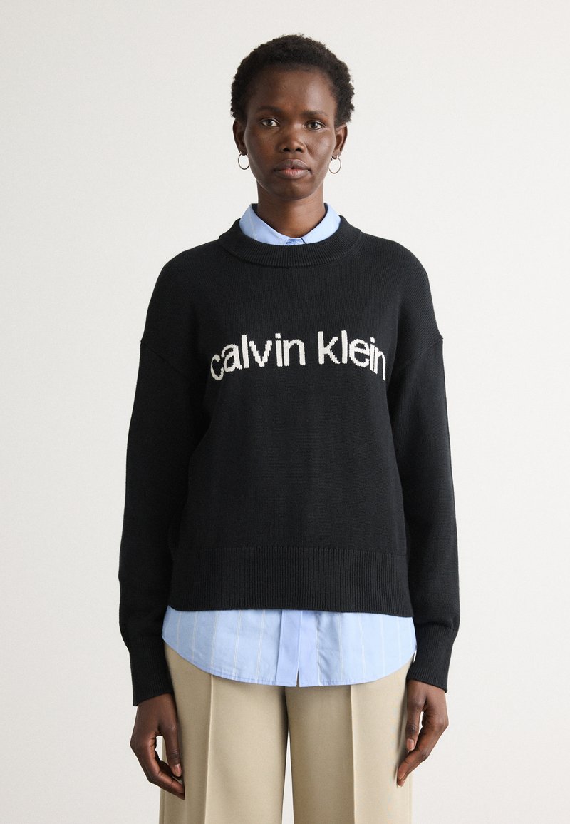 Schwarzer Strickpullover mit "Calvin Klein" Logo, Rundhalsausschnitt, überschnittenen Schultern, kombiniert mit einem hellblauen gestreiften Hemd und beigen Hosen.