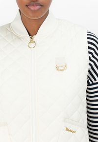 Gequilted crèmewit vest met een gouden rits, staande kraag, voorzakken en een stoffen lus; gedragen over een zwart-wit gestreept shirt.