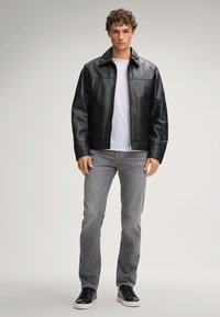 Giacca di pelle nera con collo, maniche lunghe, chiusura con zip, indossata sopra una t-shirt bianca e jeans grigi. Sneakers nere completano l'outfit.