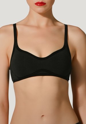 Donna che indossa un semplice reggiseno nero con spalline sottili, mostrata da sotto il mento fino alla vita su uno sfondo uniforme.