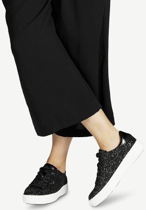 Jambes portant un pantalon large noir et des baskets noires pailletées avec des semelles blanches, un pied légèrement levé sur fond blanc.