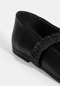 Schwarze Lederflache Schuhe mit einem gerafften Riemen über dem Obermaterial. Der Schuh hat einen niedrigen Absatz und eine strukturierte Innenauskleidung.