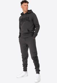 Grå hoodie og sæt med sweatpants med sorte logo detaljer. Blødt materiale, afslappet pasform. Sat sammen med hvide sneakers for et afslappet look.