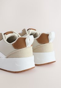 Baskets beiges avec semelles blanches, présentant une tige texturée, des accents en daim marron et des lacets contrastants. Tirette au talon visible et détails de couture.