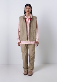 Beige vest med glidelås og sidelommer over en lyserosa skjorte. Løse beige bukser og brune sko fullfører antrekket.