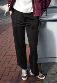 Pantalons noirs à jambes larges avec une texture lisse, dotés de poches avant et de plis marqués, associés à des baskets blanches et bordeaux.