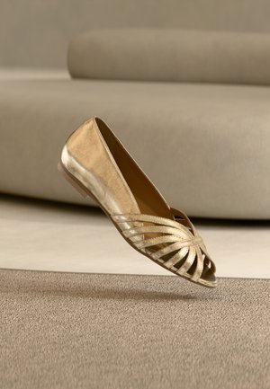 Gouden metallic platte schoen met open teen en kruisbandontwerp, geplaatst boven een beige tapijt en voor een taupe bank.