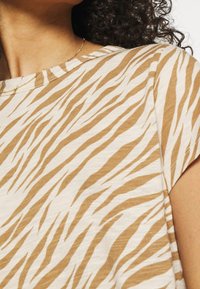 Beige topp med en krämfärgad bas som har ett vågigt randmönster i en varm brun nyans. Korta ärmar och rund halsringning. Mjuk tygkvalitet.