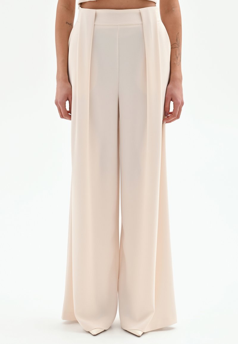 Pantalones de pierna ancha en beige claro, con cintura alta, textura suave y sutiles detalles de drapeado. Ajuste estructurado, diseño fluido.