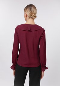 Burgundy blouse with a ruffled collar and long sleeves. Textured fabric with gathered cuffs and a relaxed fit. Black pants not included.