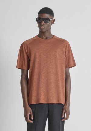 Uomo con camicia a maniche corte color ruggine oversize e pantaloni neri, accessoriate con grandi occhiali da sole neri, in piedi davanti a uno sfondo grigio.